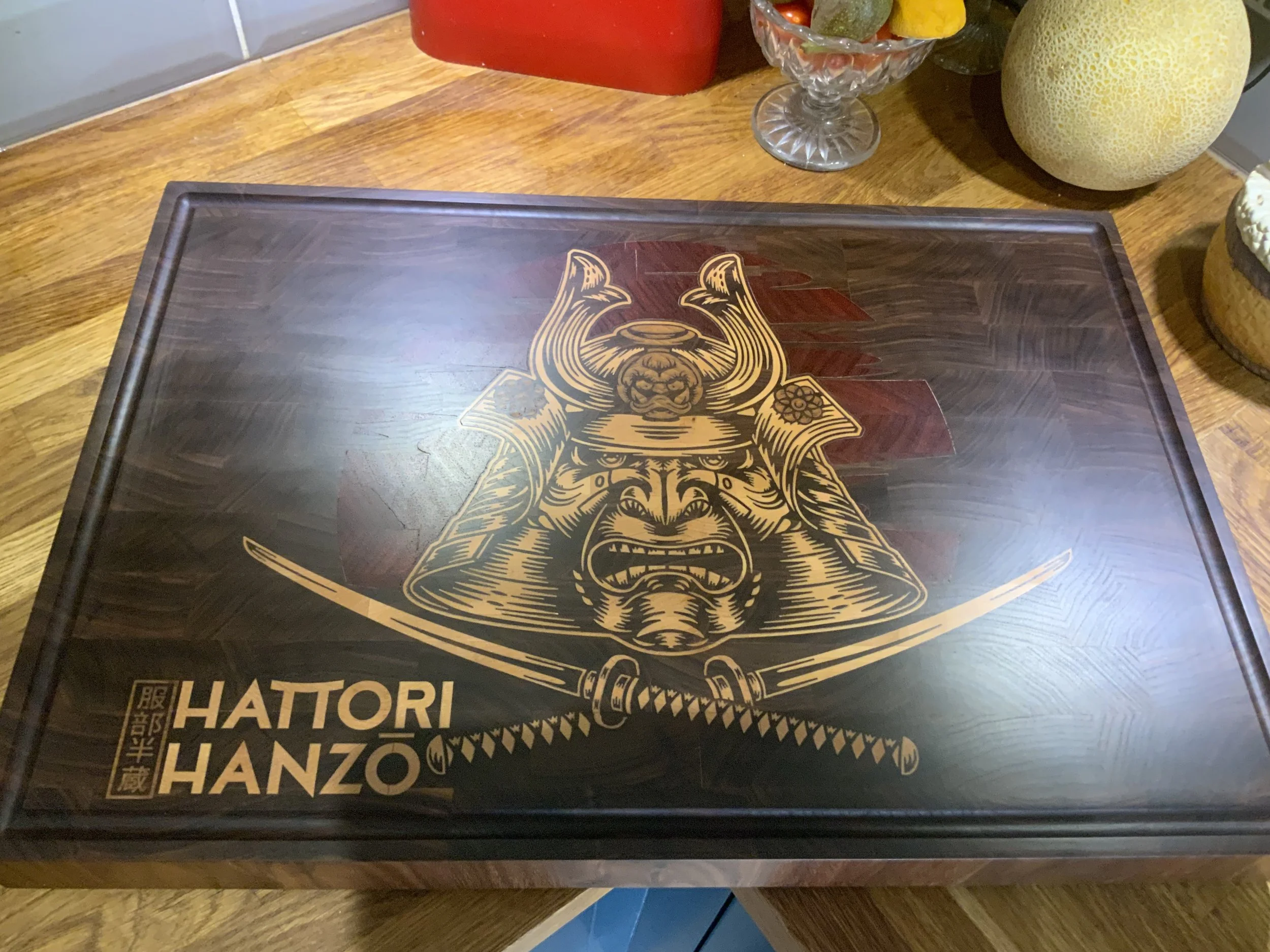 A wooden table featuring a decorative box with a gold samurai helmet and face design, the words "HATTORI HANZO" and Japanese characters printed on it. In the background, there are decorative items including a red container, a glass bowl with fruit, and some round decorative objects.