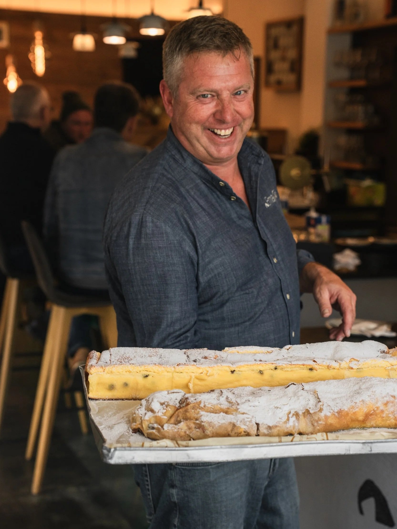 Ein lachender Mann hält ein großes Stück Kuchen in einer Bäckerei oder einem Café.