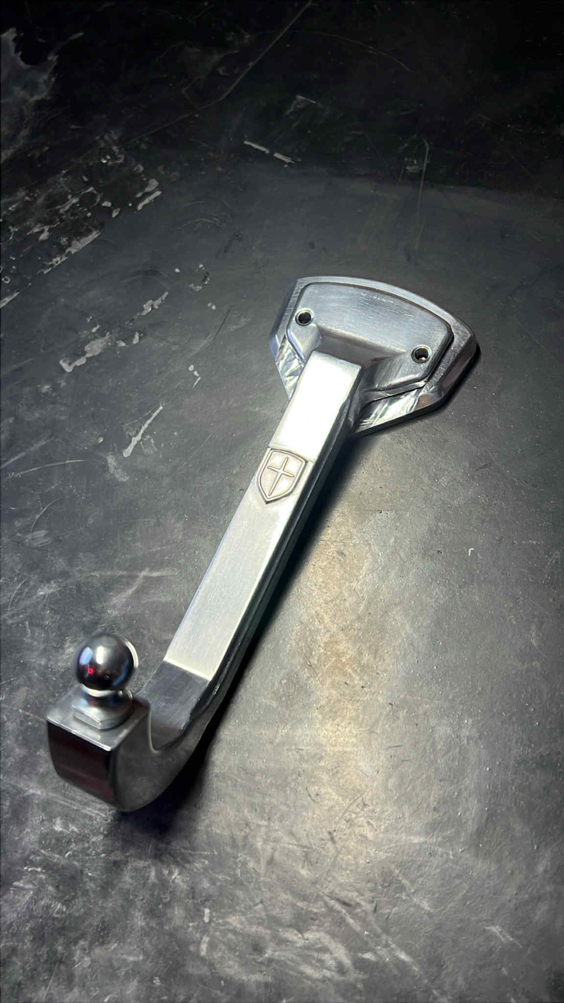 A metallic trailer hitch with a ball and mounting plate lying on a dark textured surface.