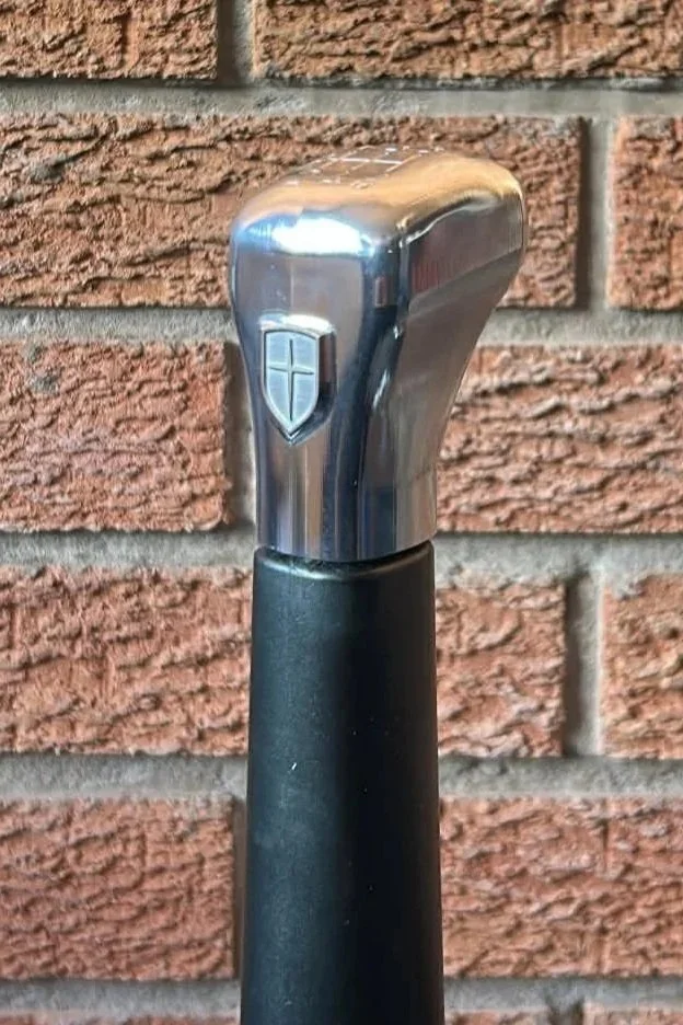 A close-up of a metal can opener handle with a shield icon, mounted on a black handle, against a brick wall background.