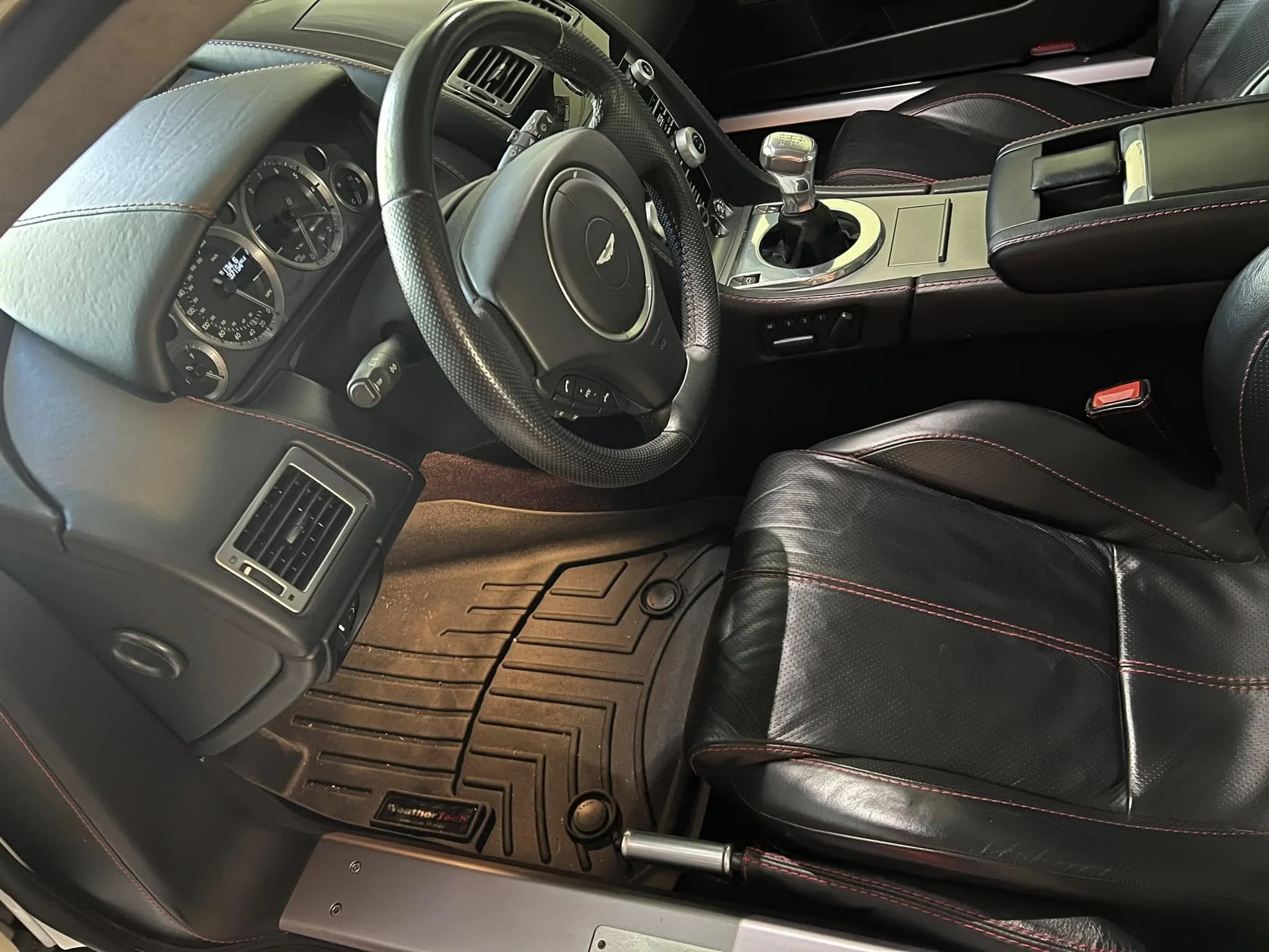 Interior of a sports car with a black leather seat with red stitching, a gear shifter, dashboard gauges, air vents, and floor mats.