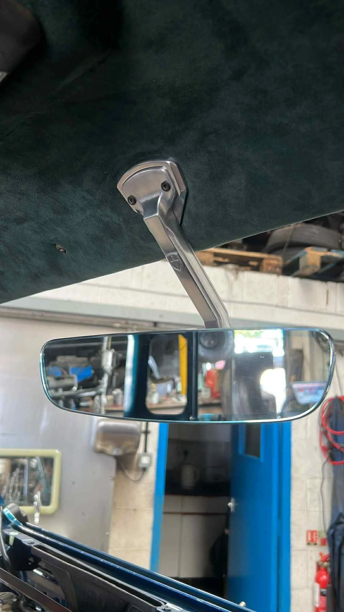 A rearview mirror attached to a car's ceiling inside a garage, with workshop tools and equipment visible in the background.