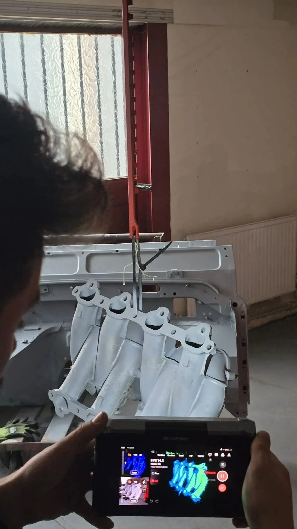 A person is filming an engine block hanging from a red hoist in a workshop, with a window and a radiator in the background.