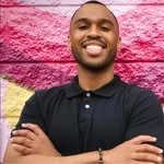 A smiling man with short hair and a beard stands in front of a colorful pink and purple background, wearing a black polo shirt.