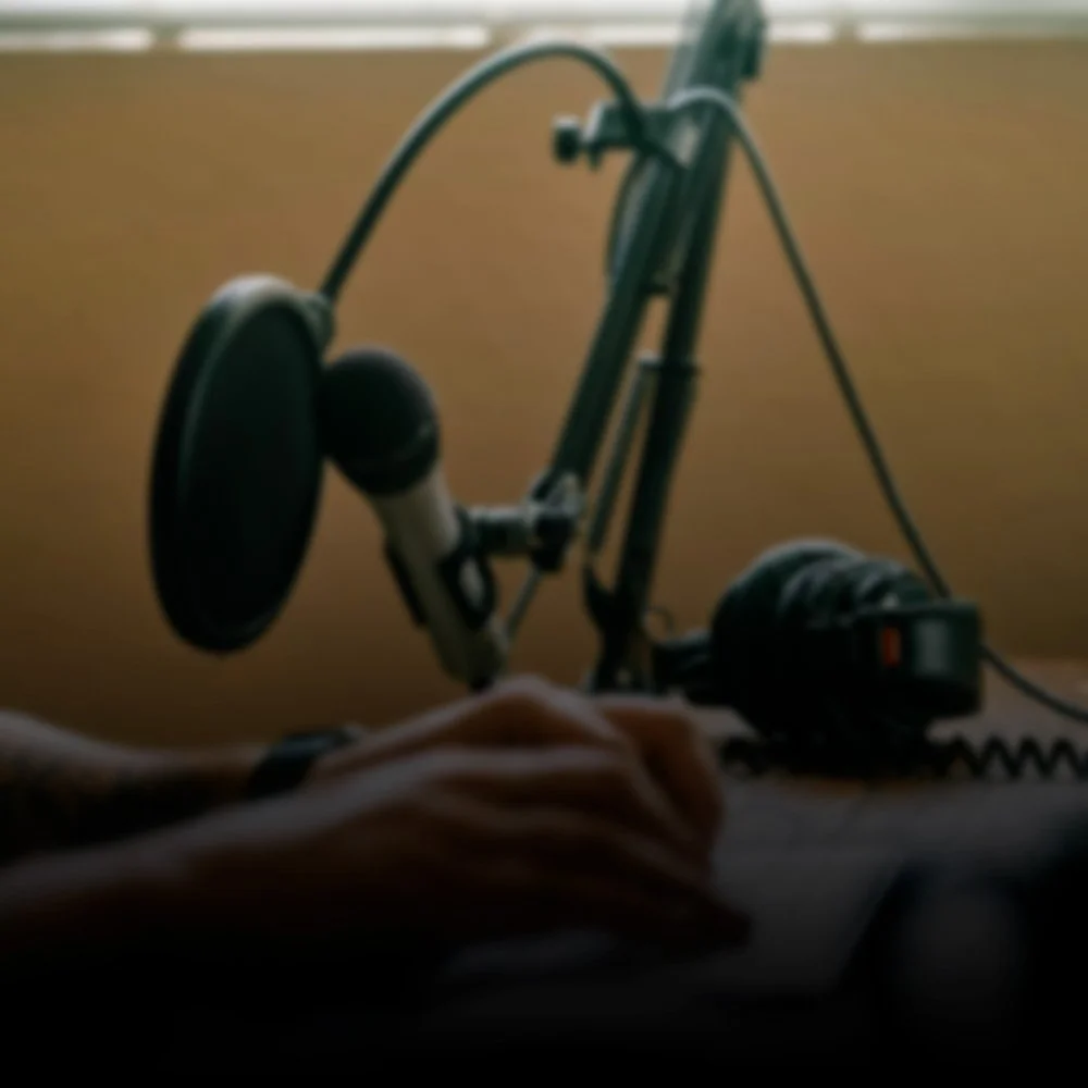 Close-up of a microphone, headphones, and a person's hand on a computer mouse on a desk, with a brown wall in the background.