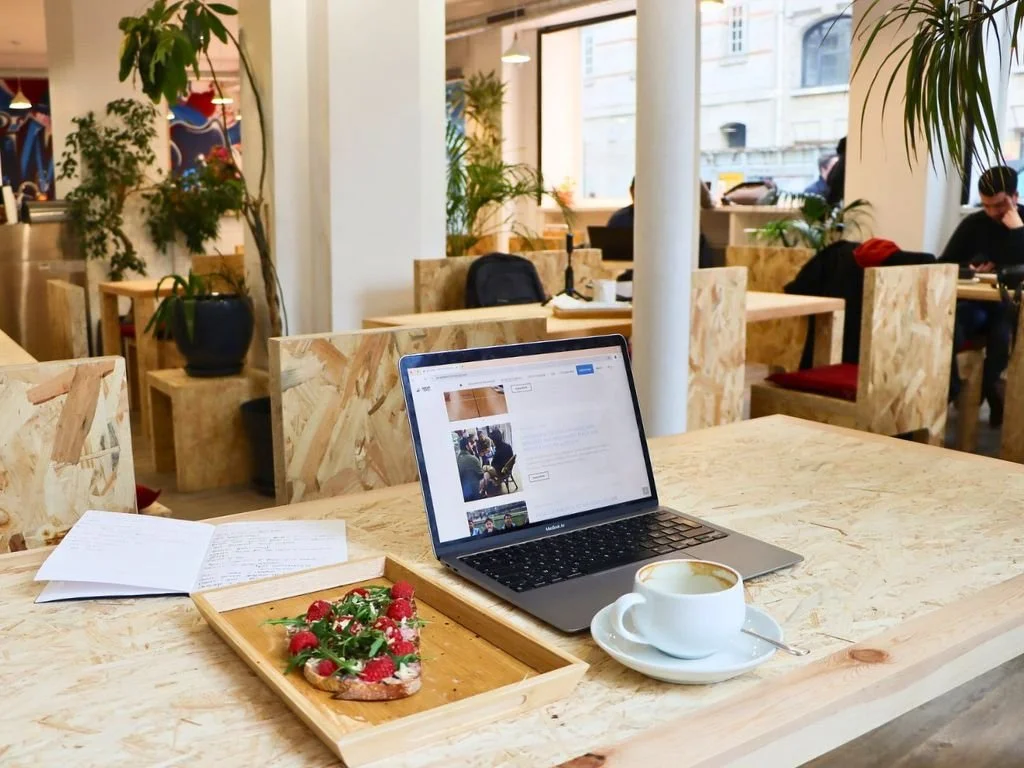 Laptop open on a wooden table with a notebook, a slice of berry-topped cake, and a cup of coffee in a cafe with plants and other customers in the background.