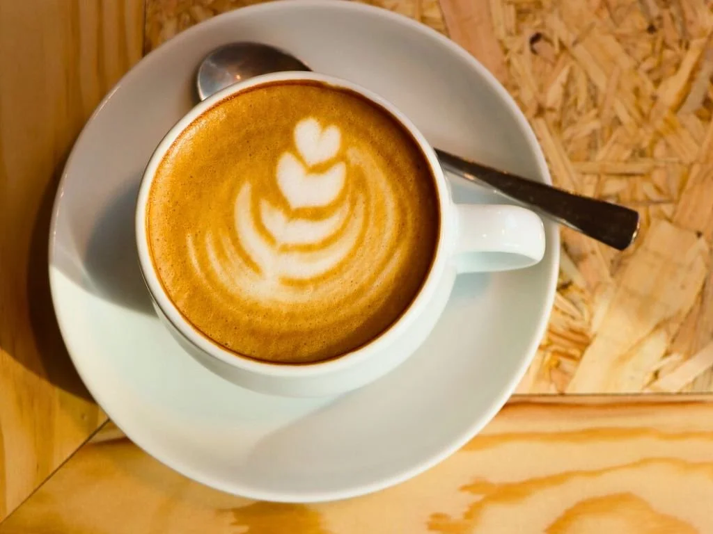 A cup of coffee with latte art on top, served on a white saucer with a spoon, on a wooden table.