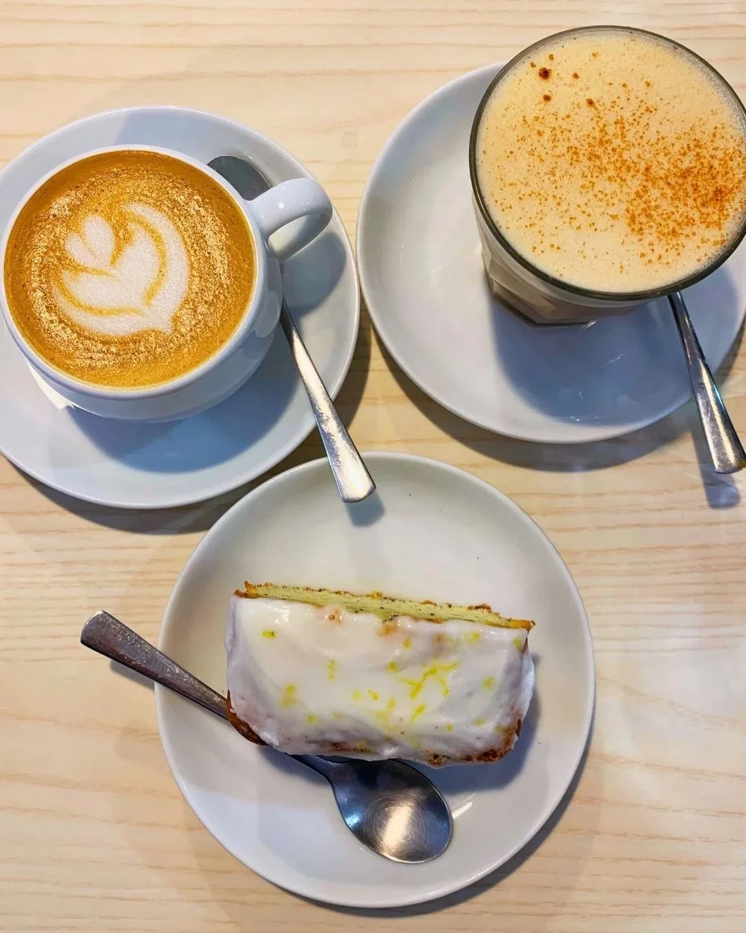 A table with two cups of coffee, a glass of a frothy drink, and a slice of cake with white icing.