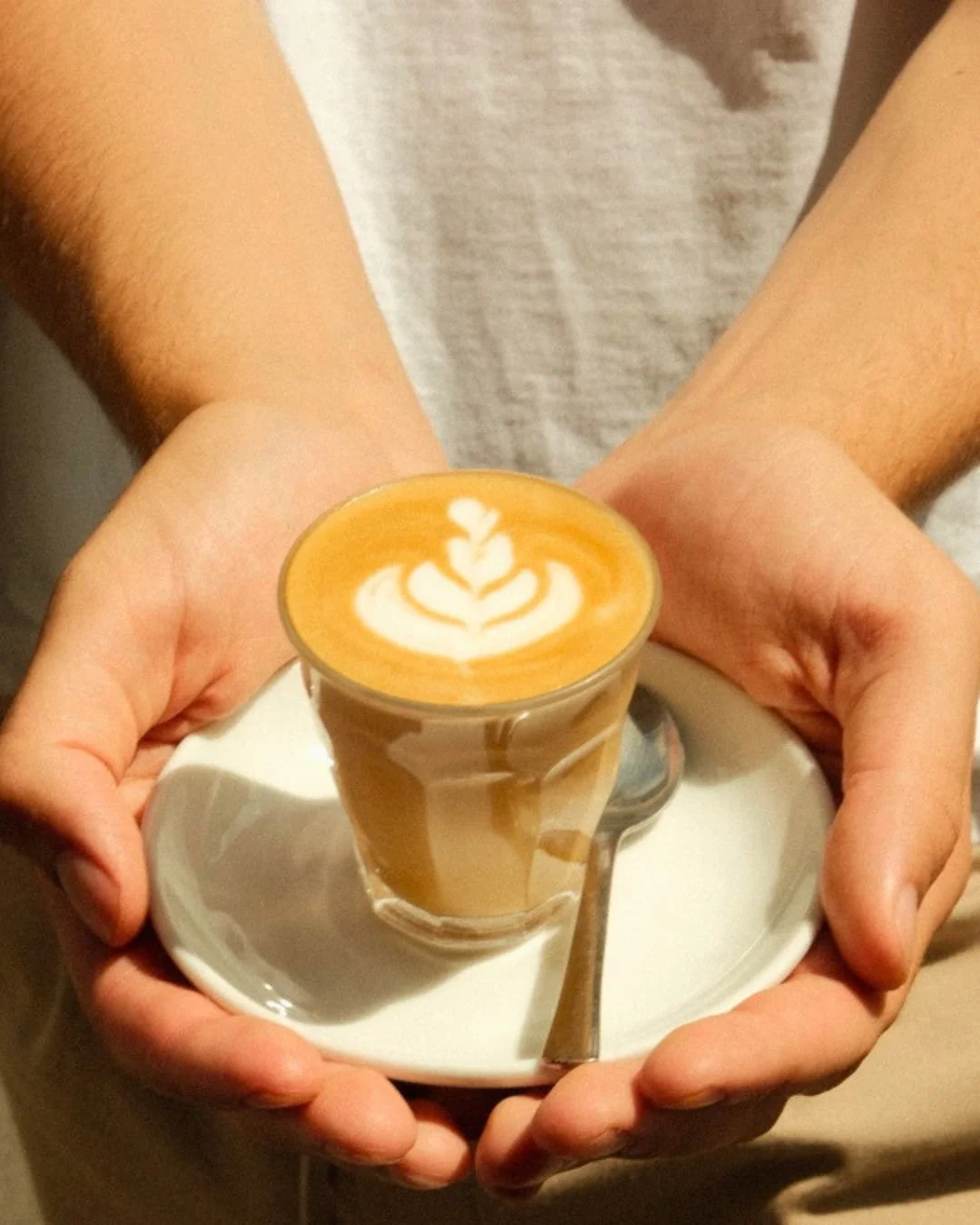 A person holding a white plate with a small glass of latte art coffee on it, featuring a leaf design, with a spoon resting on the plate.