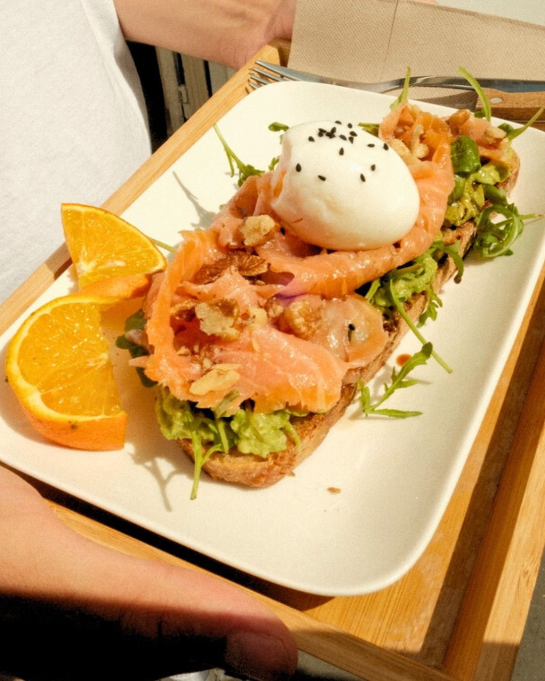 Open-faced sandwich with smoked salmon, leafy greens, a poached egg topped with black sesame seeds, and orange slices on a white rectangular plate.