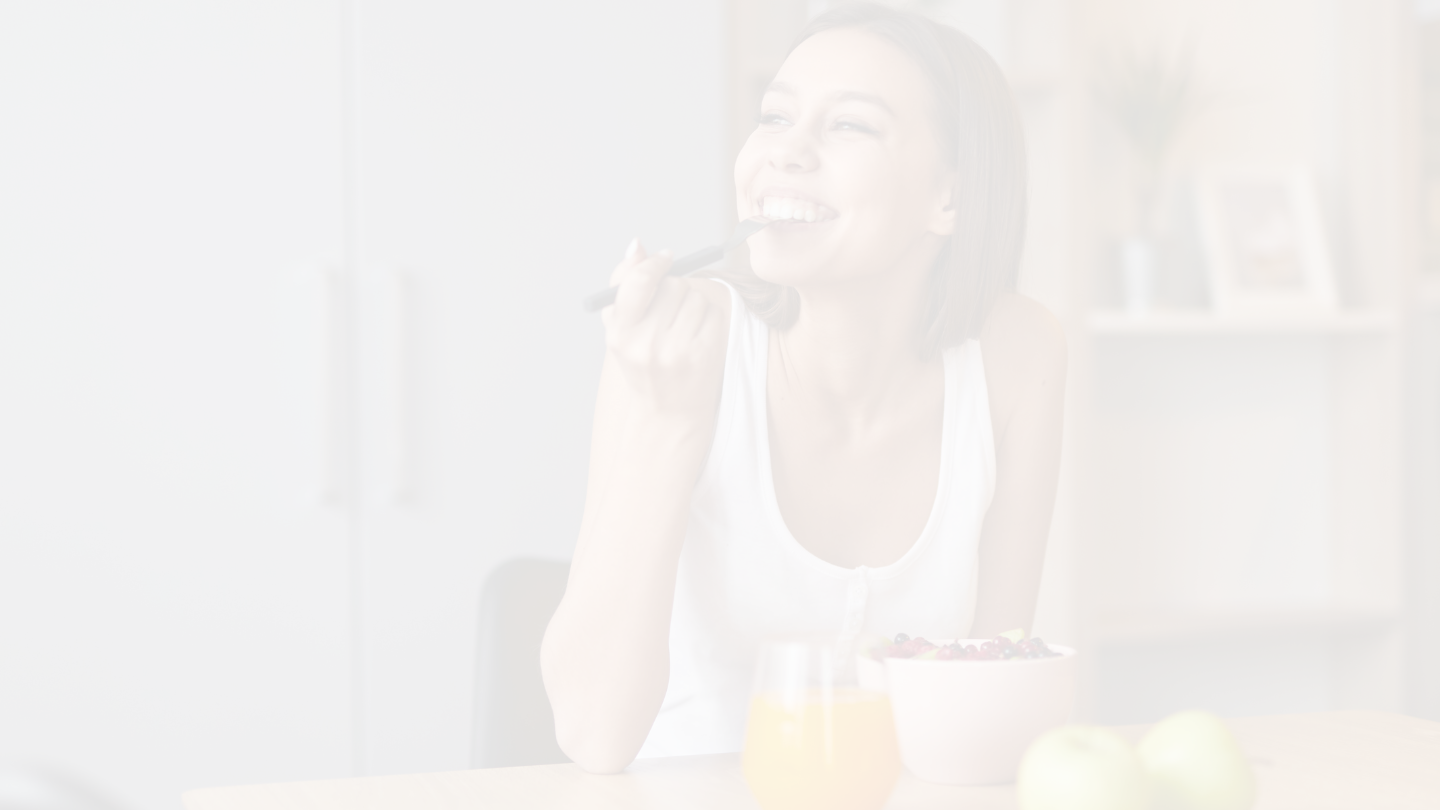 A woman smiling and eating with a spoon at a table with a bowl of fruit and a glass of orange juice.