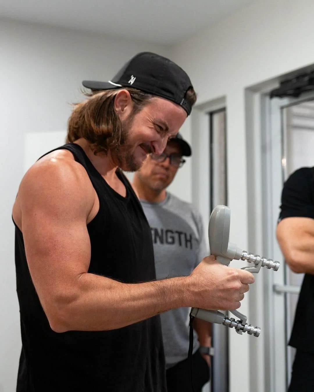 A man with long hair, wearing a black sleeveless shirt and a backward cap, smiling while holding a massage gun. Other people are in the background.