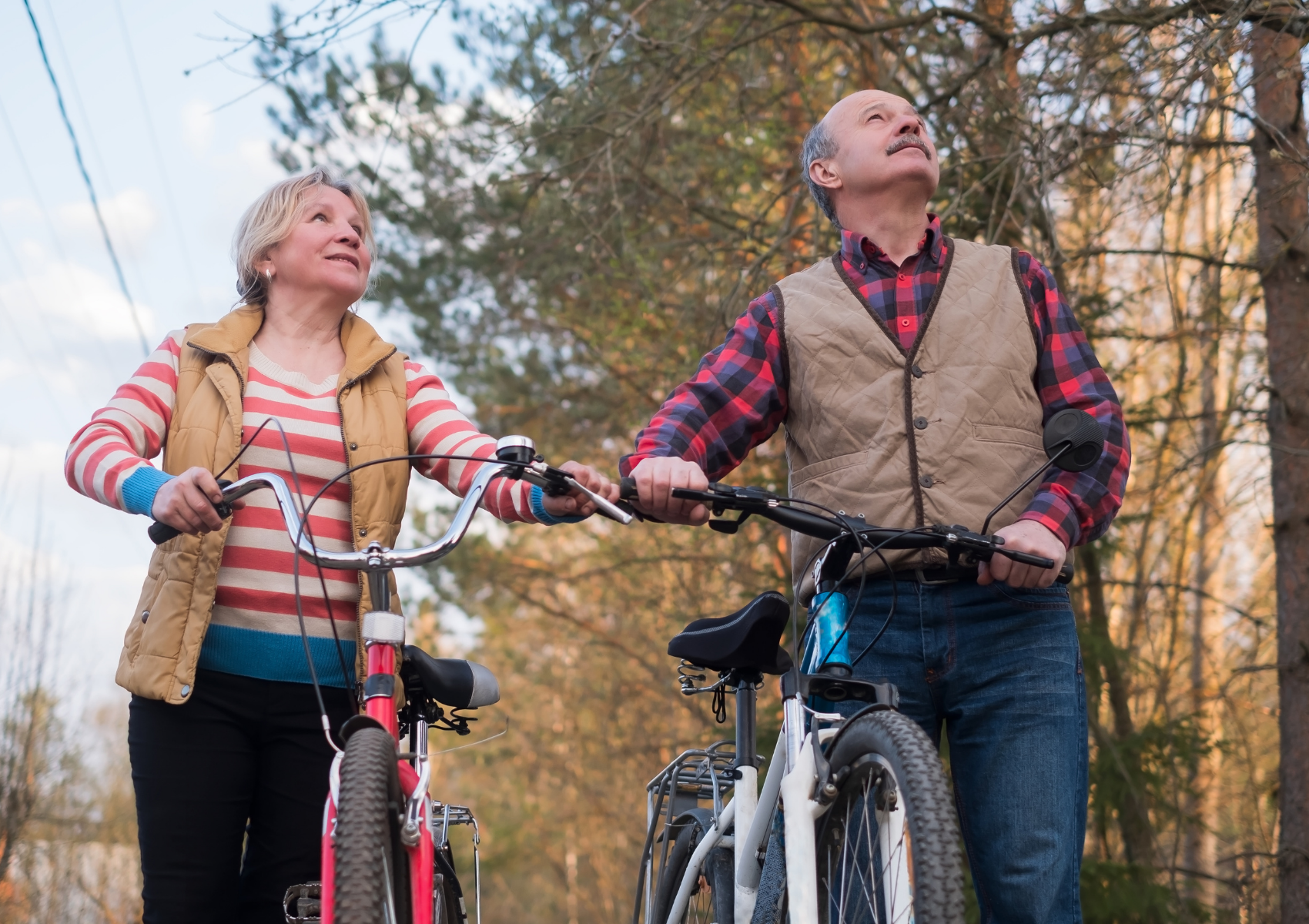 An elderly couple with bicycles look up at the sky during a fall day with colorful trees