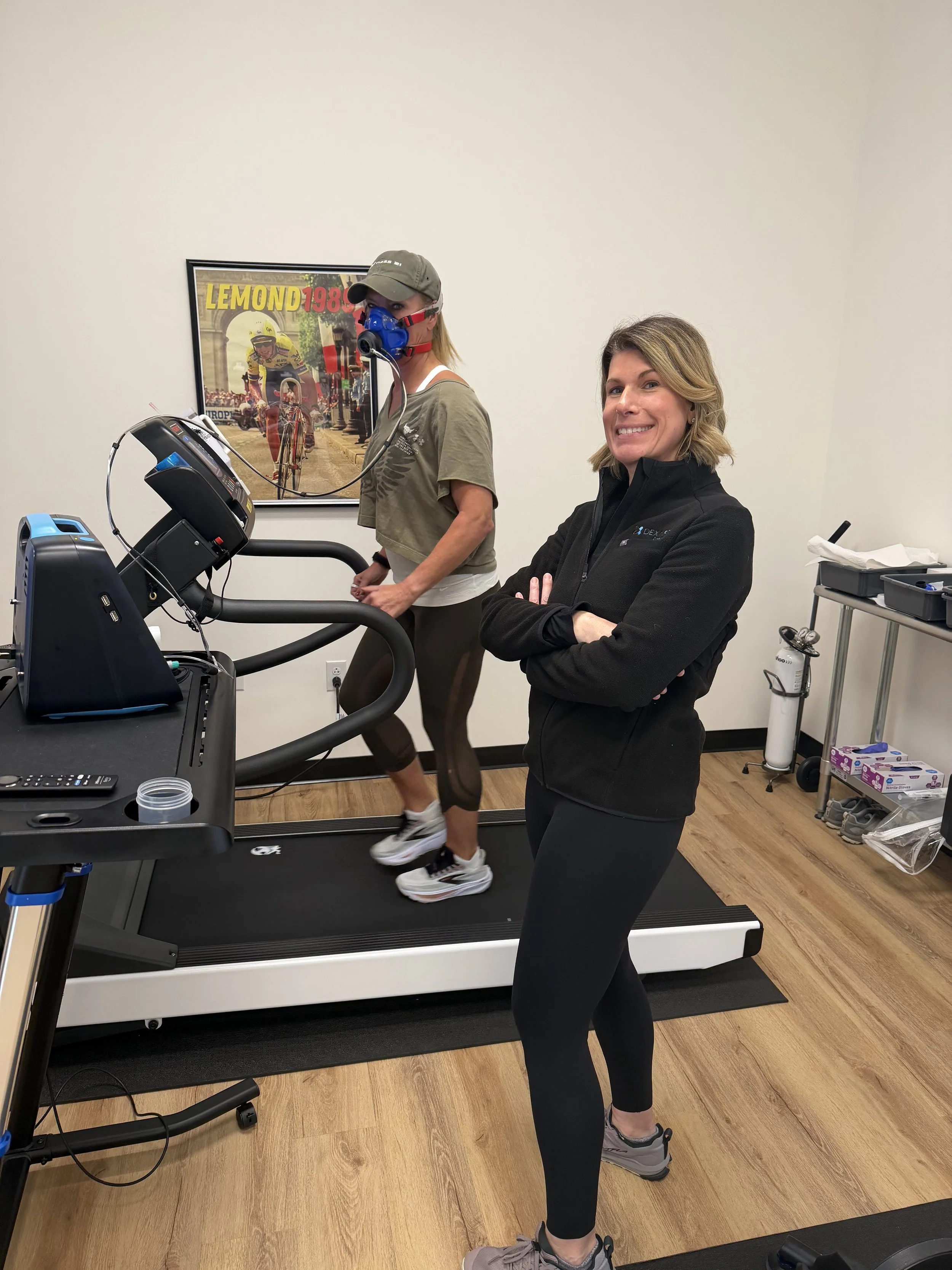 Woman on a treadmill in a gym, wearing black leggings, gray shoes, and a black jacket, with her arms crossed, smiling at the camera. Behind her, another woman wearing a mask, cap, and casual clothes uses the treadmill.