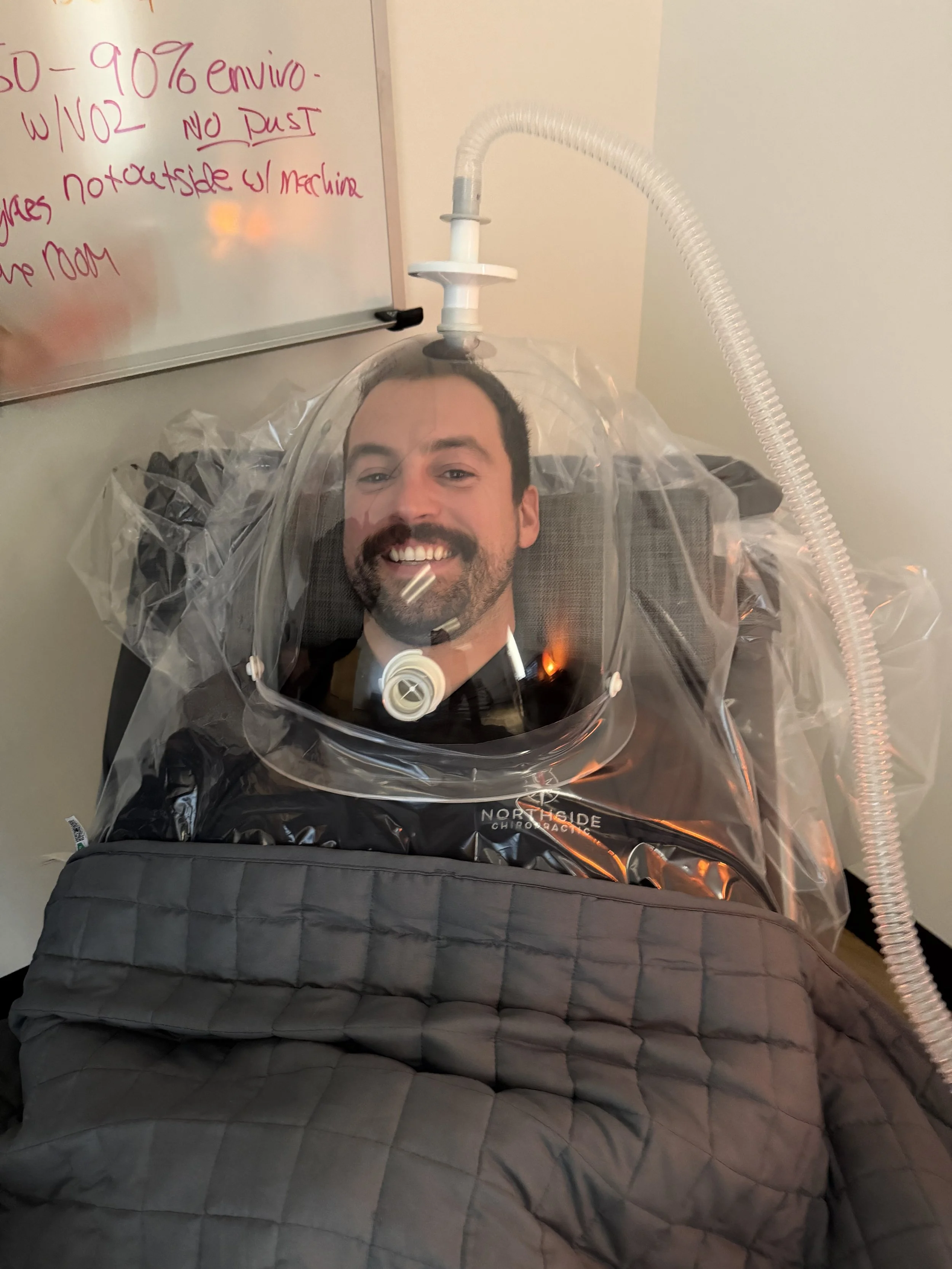 A man laying back in a chair with a clear canopy connected to a breathing tube, smiling at the camera completing a resting metabolic rate test in DexaFit Greenville office.