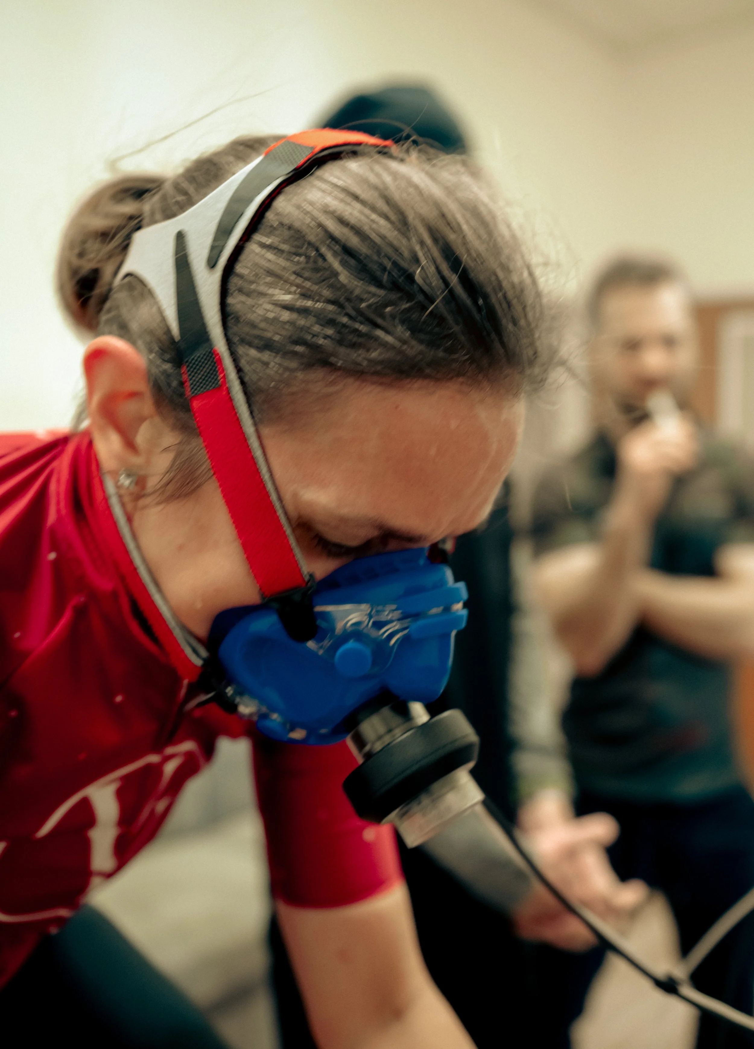 Woman with blue mask and red head strap with wires leading to metabolic cart. She is performing VO2 Test. Man in background observing.