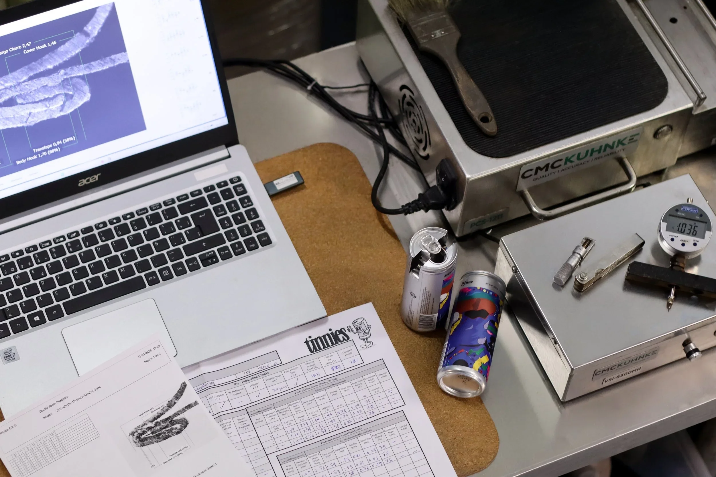 Laboratory or quality control workspace with a laptop displaying a graph, printed documents, a small digital scale showing 410.36, two cans of energy drinks, a metal tray with tools including a wrench and a screwdriver, and a portable burner or heating device.