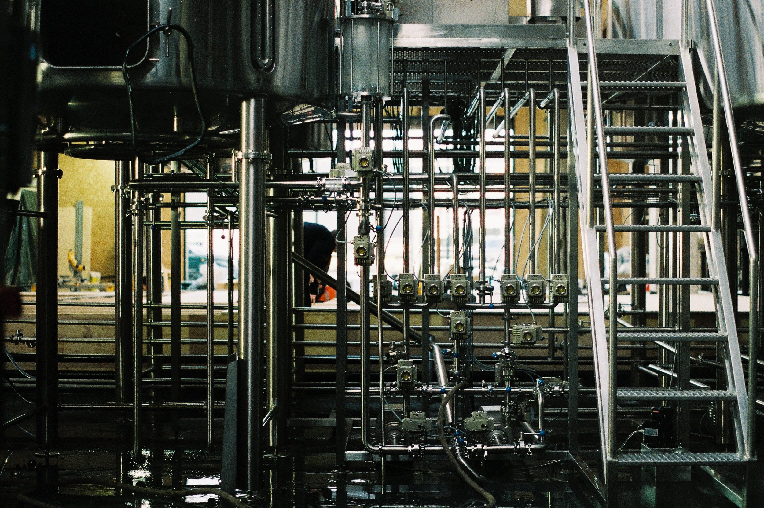 Industrial stainless steel equipment with pipes, valves, and a metal staircase in a factory setting.