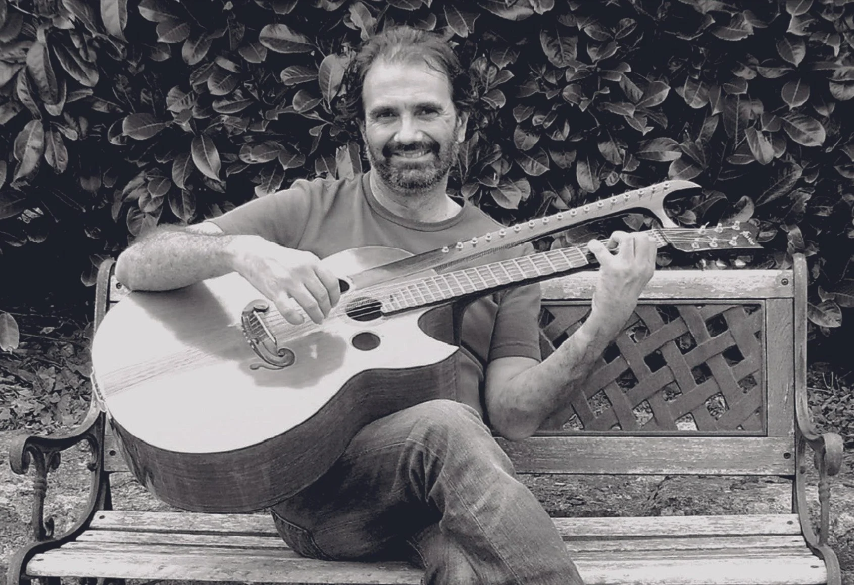 Homme assis sur un banc en bois, jouant de la guitare, avec un arrière-plan de feuilles d'arbres.