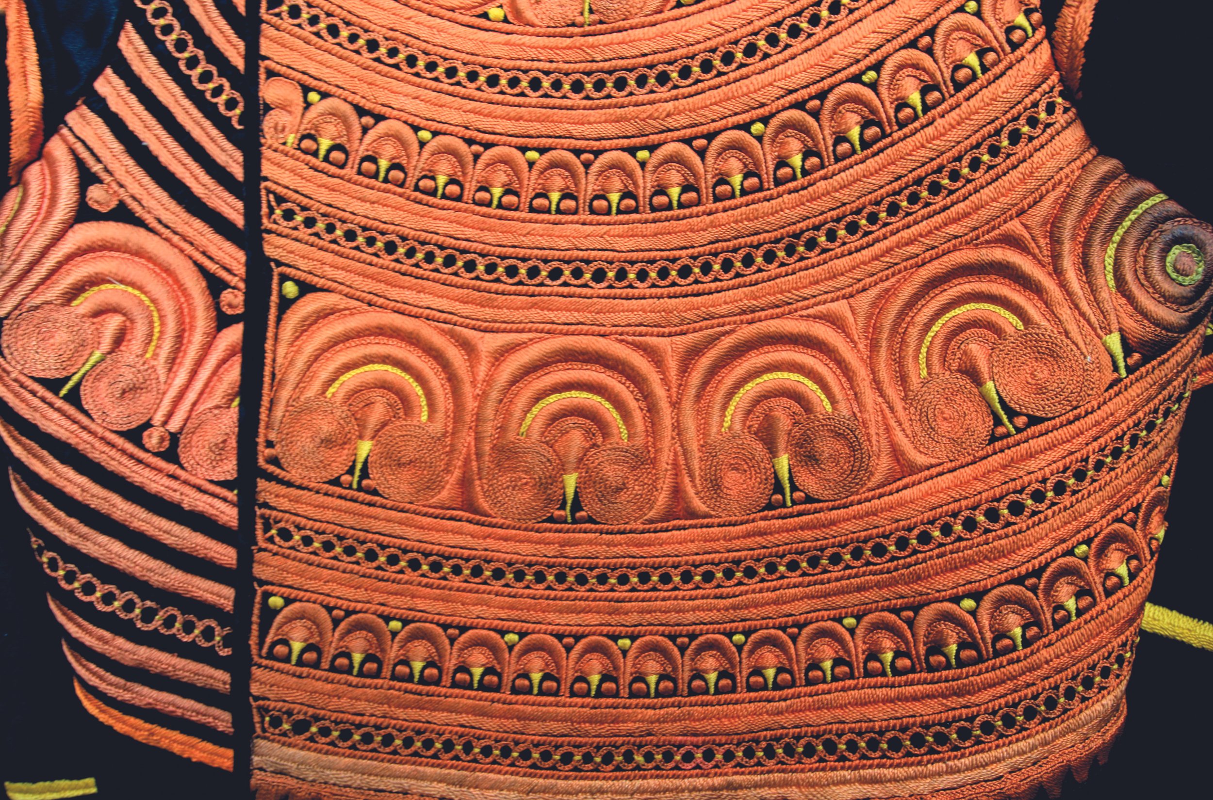 Détail d'une broderie traditionnelle orange avec des motifs en spiral et en arc, accentuée par des broderies noires et jaunes.