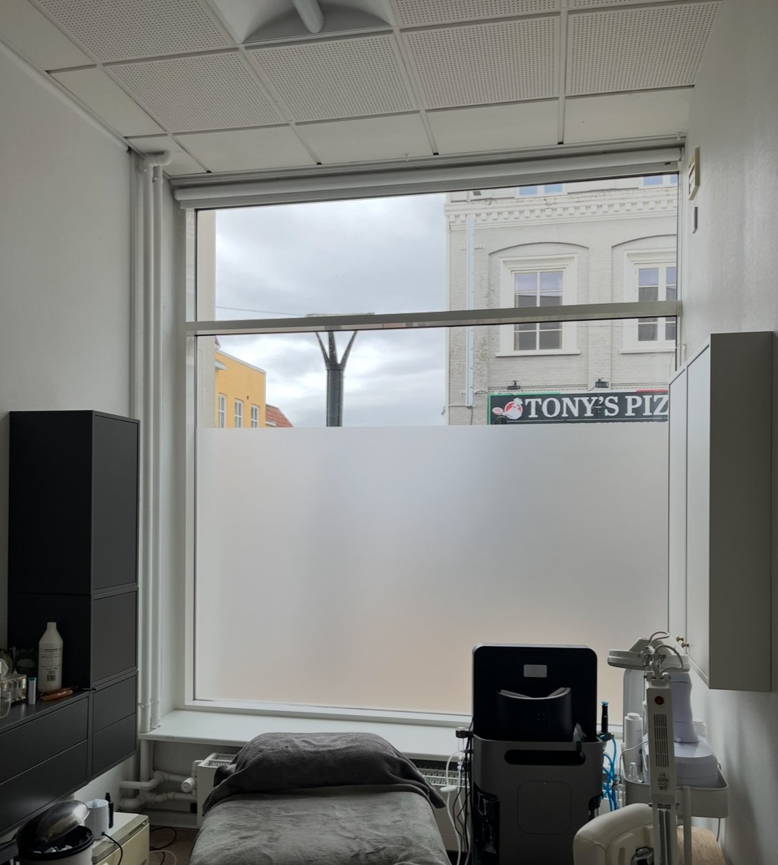 A medical or treatment room with a bed, cabinets, and medical equipment, with a large window showing a cloudy sky and a building with a sign that reads "Tony's Piz" outside.
