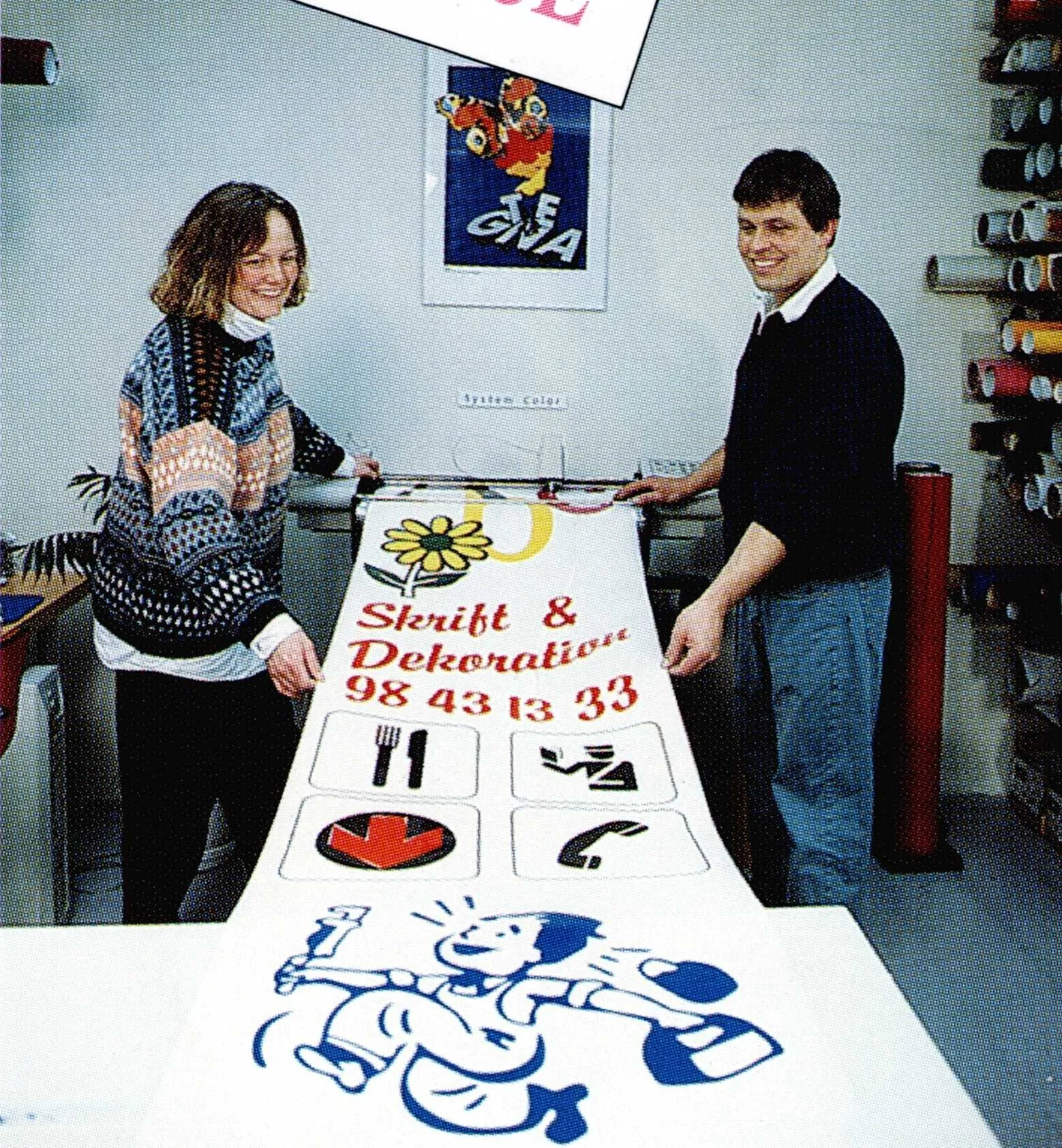 Two people holding a decorated cloth with symbols, a phone, and a phone receiver, in a room with a poster of a cartoon duck on the wall and shelves of rolls of material.