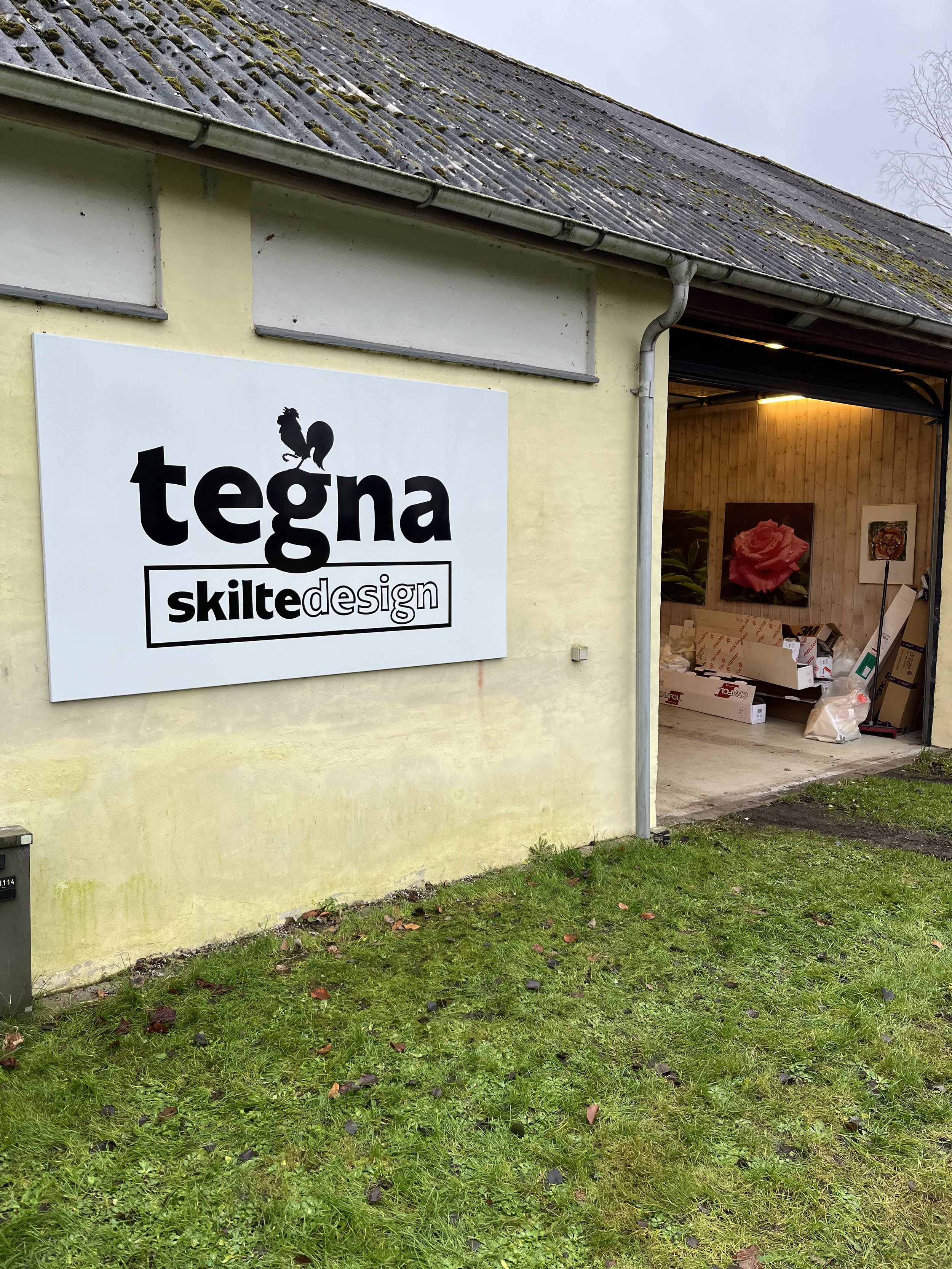 Sign on a yellow building that reads 'Tegna Skilte Design' with a logo of a rooster on top of the text. The building has a garage door open to the interior, revealing framed pictures and supplies.