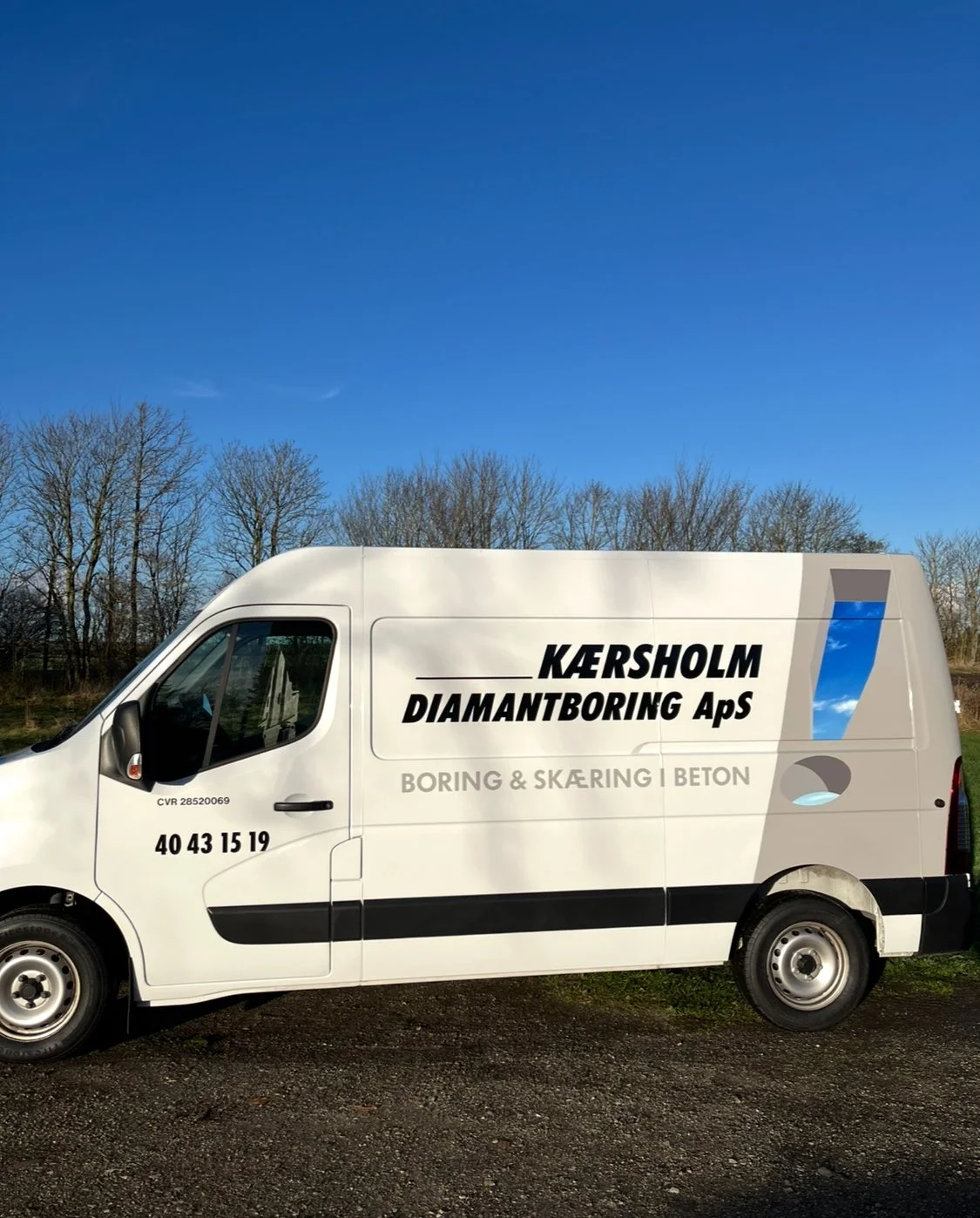 A white work van parked outdoors on a gravel surface, with a background of leafless trees and a clear blue sky. The van has black and gray text and graphics, including company branding and contact information.
