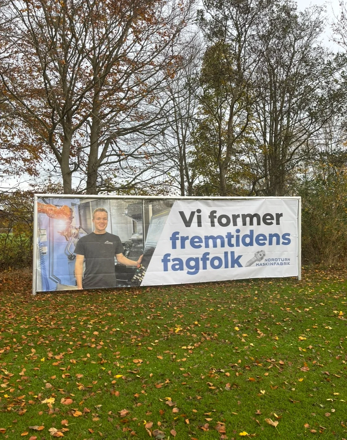 Outdoor banner promoting a technical training or craftsmanship, showing a smiling man in a black t-shirt working with machinery, with fall leaves on the grass and leafless trees in the background.