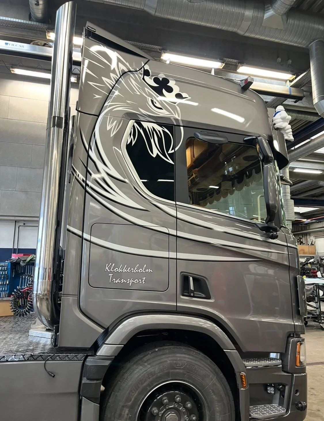 A large gray semi-truck with decorative graphics of a wolf's head and a three-leaf clover on the side, parked inside an industrial garage.