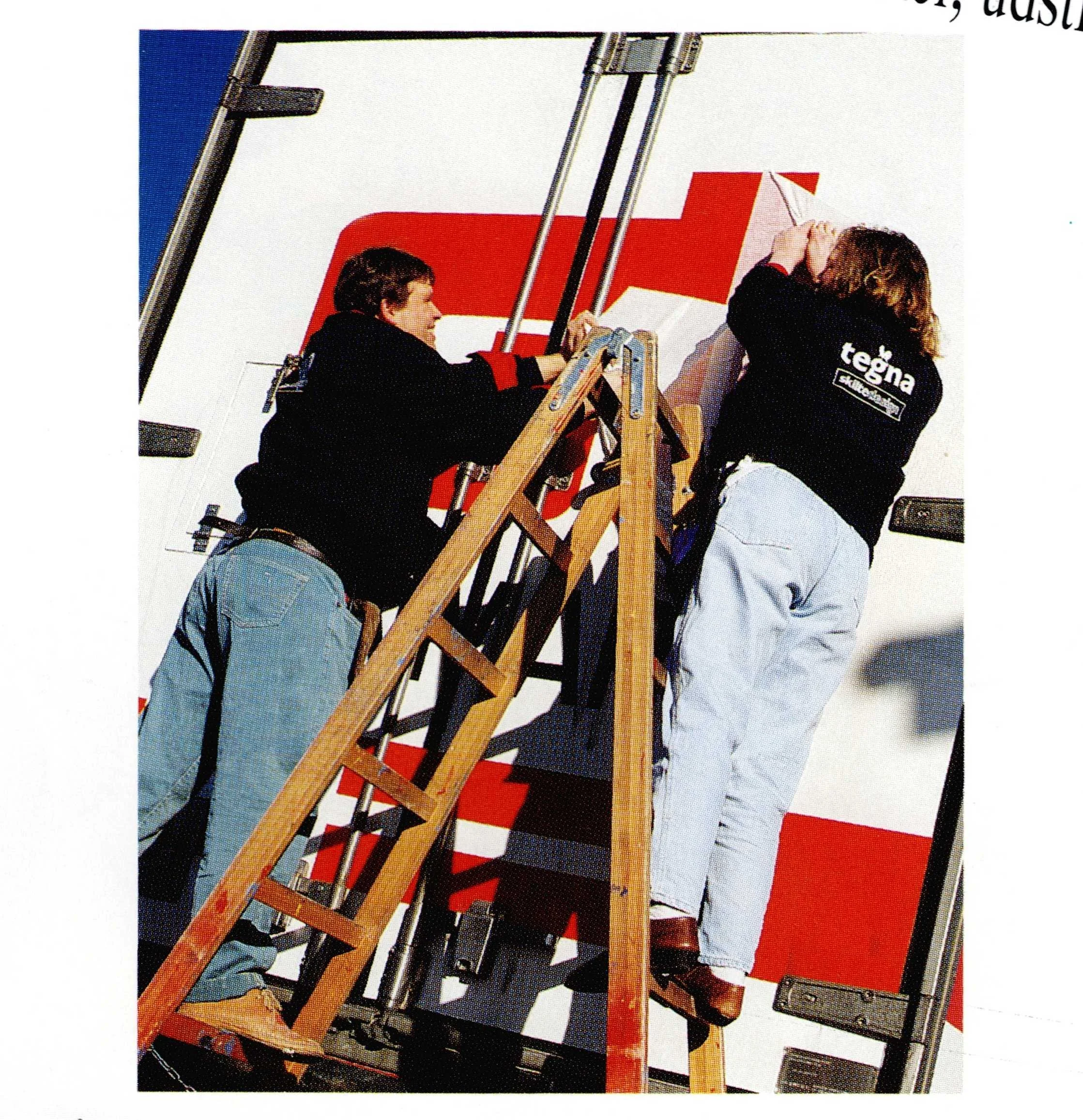 People working on a large poster or billboard, one person on a ladder and two others assisting, with a red and white graphic background.