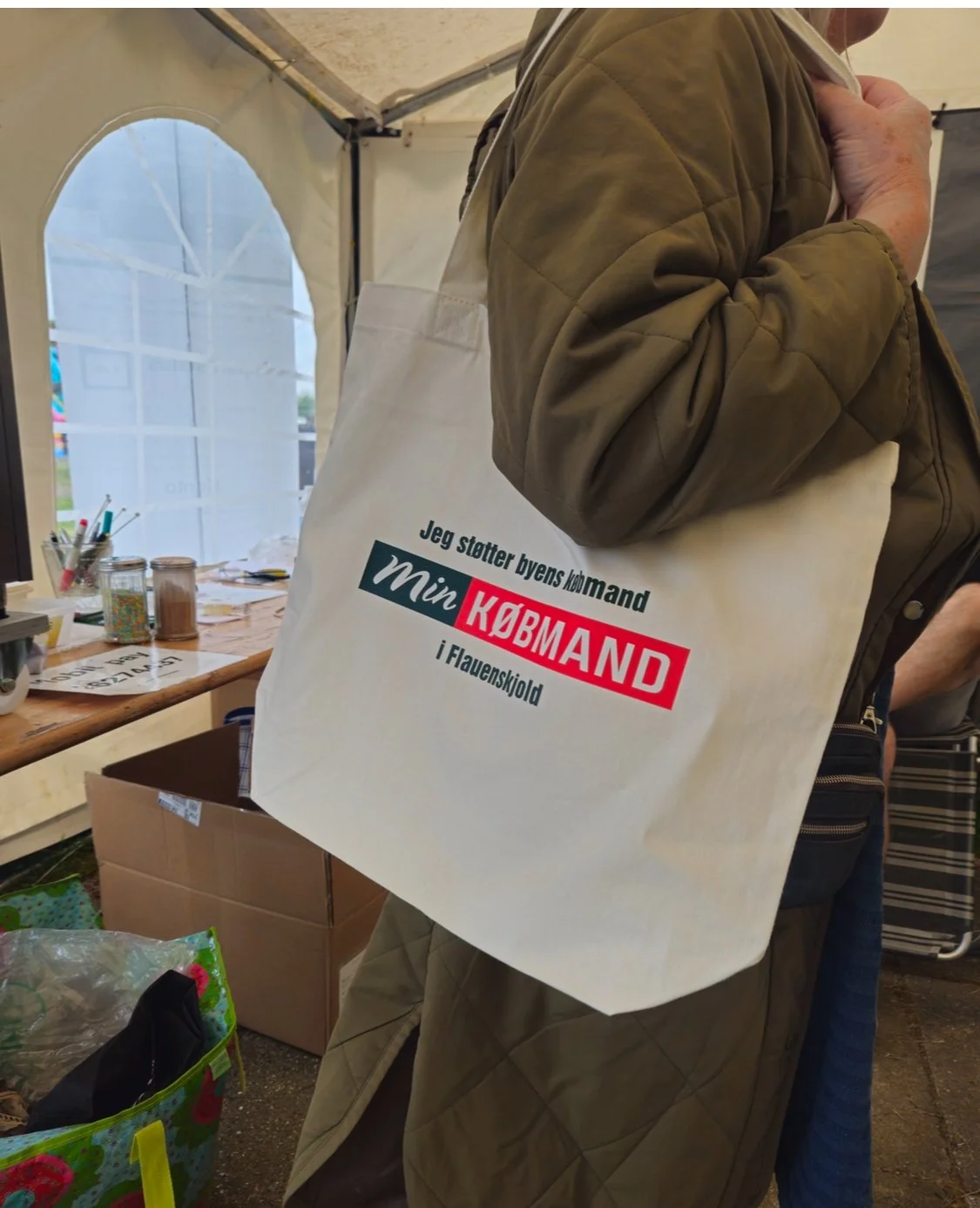 Person holding a beige tote bag with Danish text in a market or fair setting.