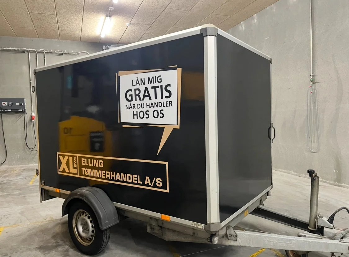 A black enclosed trailer in an indoor parking lot with a promotional sign in Norwegian advertising free loans for handling at a hardware store.