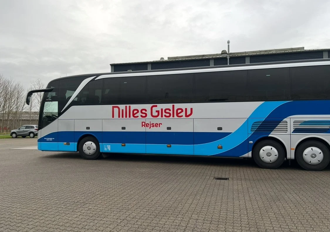 A blue and white bus with the words 'Nilles Gislev Rejser' written on the side, parked on a paved area under a cloudy sky.