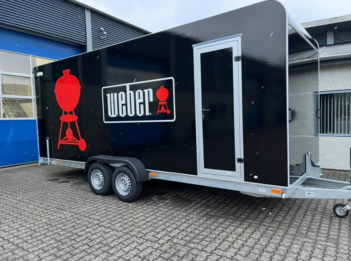 Black Weber grill-themed trailer with the Weber logo and red grill icons on the side, parked on a paved area outside a building.