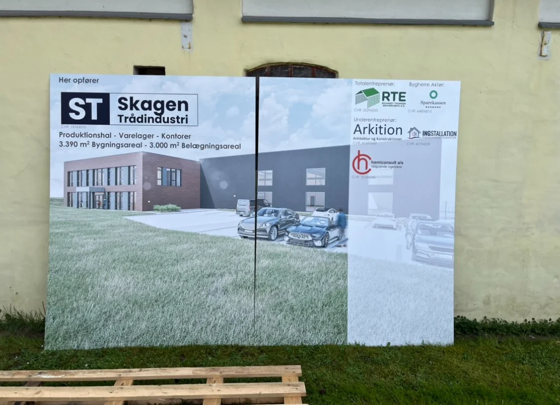 An outdoor signboard showing a construction project for a new industrial building, with parking spaces and cars in front, and various company logos and project details.