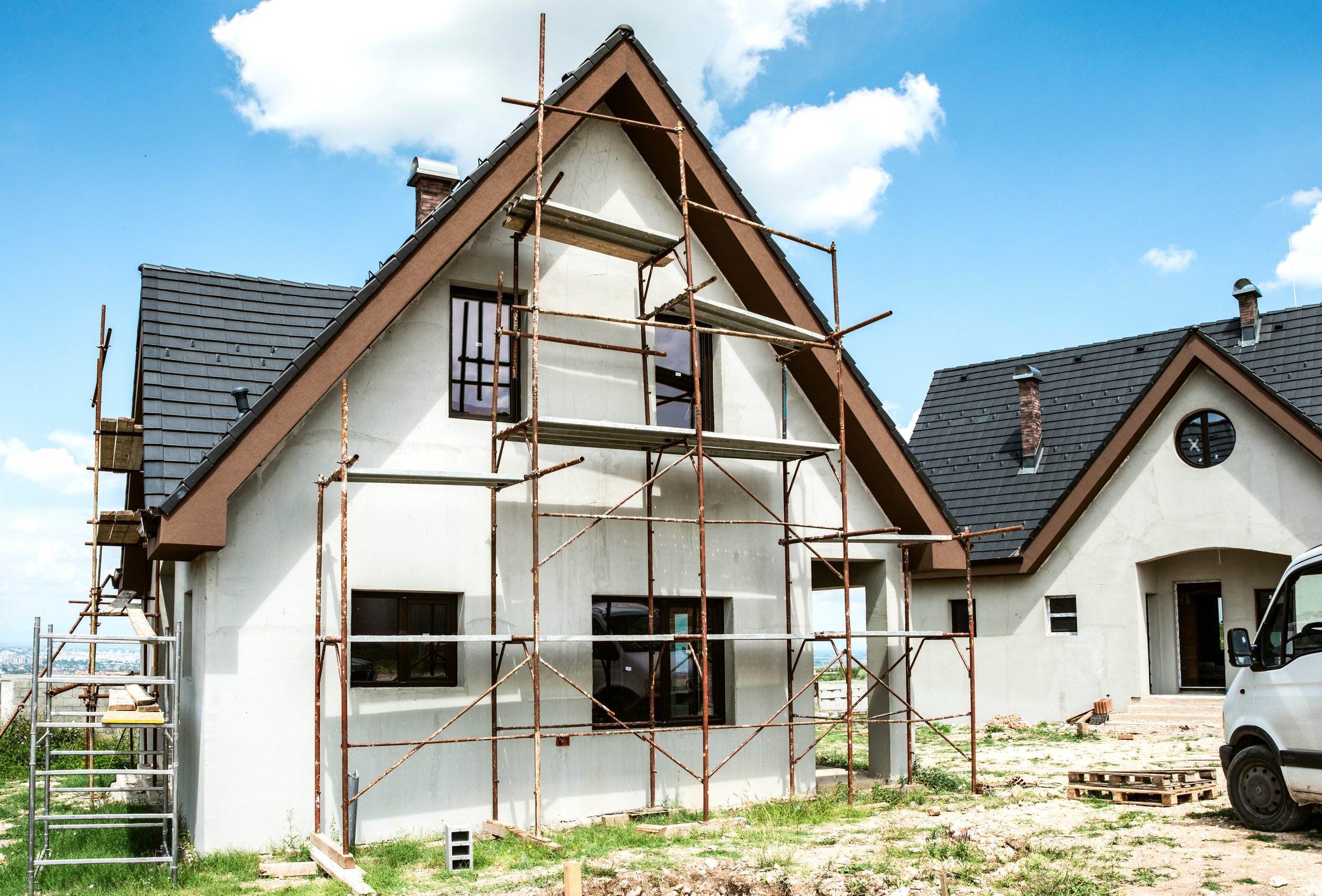 Une maison en construction avec des échafaudages en métal, la façade en cours de finition, un ciel bleu avec quelques nuages.
