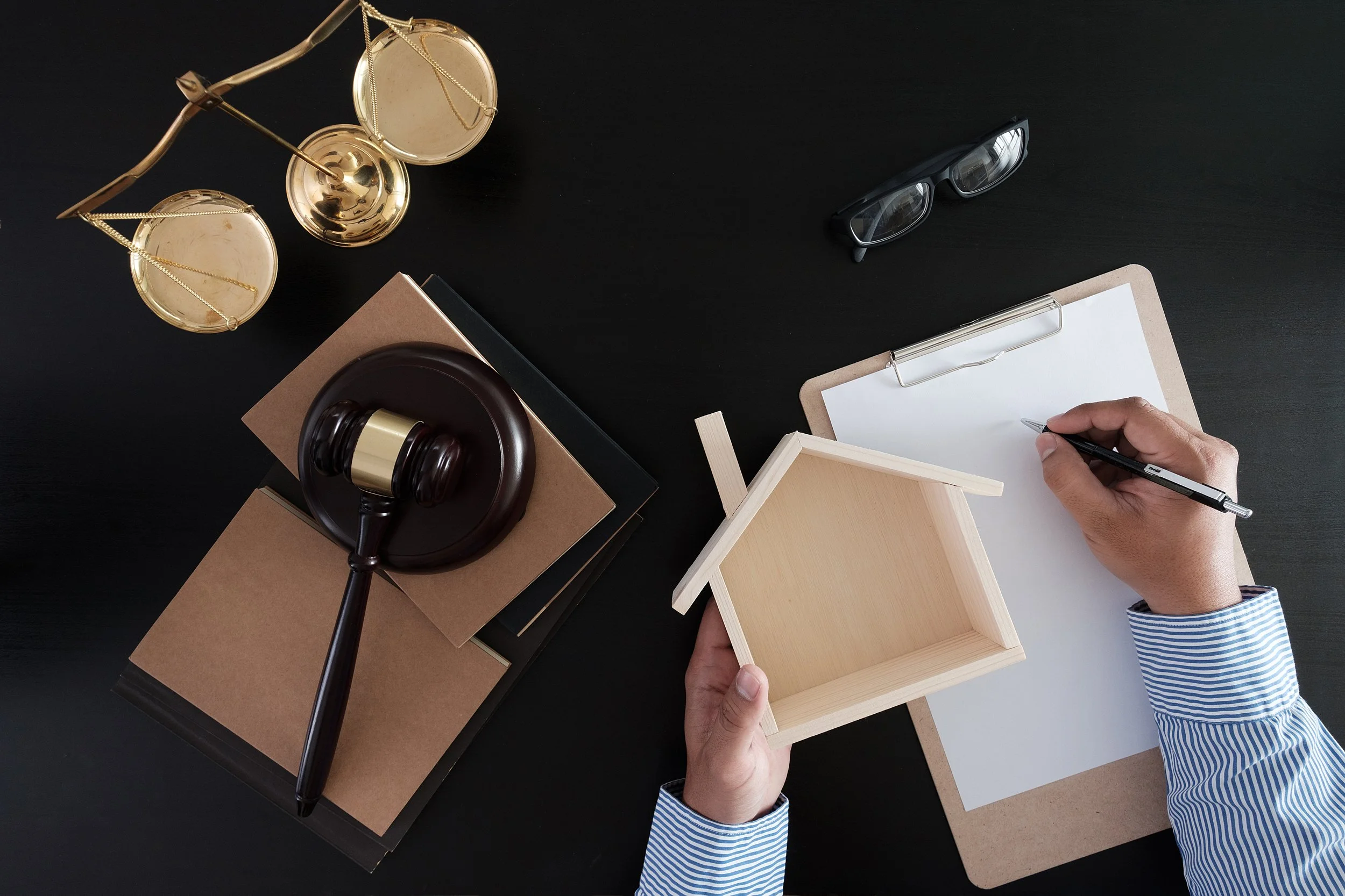 Une main tient une petite maison en bois, une autre main écrit sur un tableau blanc, à côté se trouvent une paire de lunettes, une balance de justice dorée, un marteau en bois sur un tas de livres, et un petit tableau noir. 