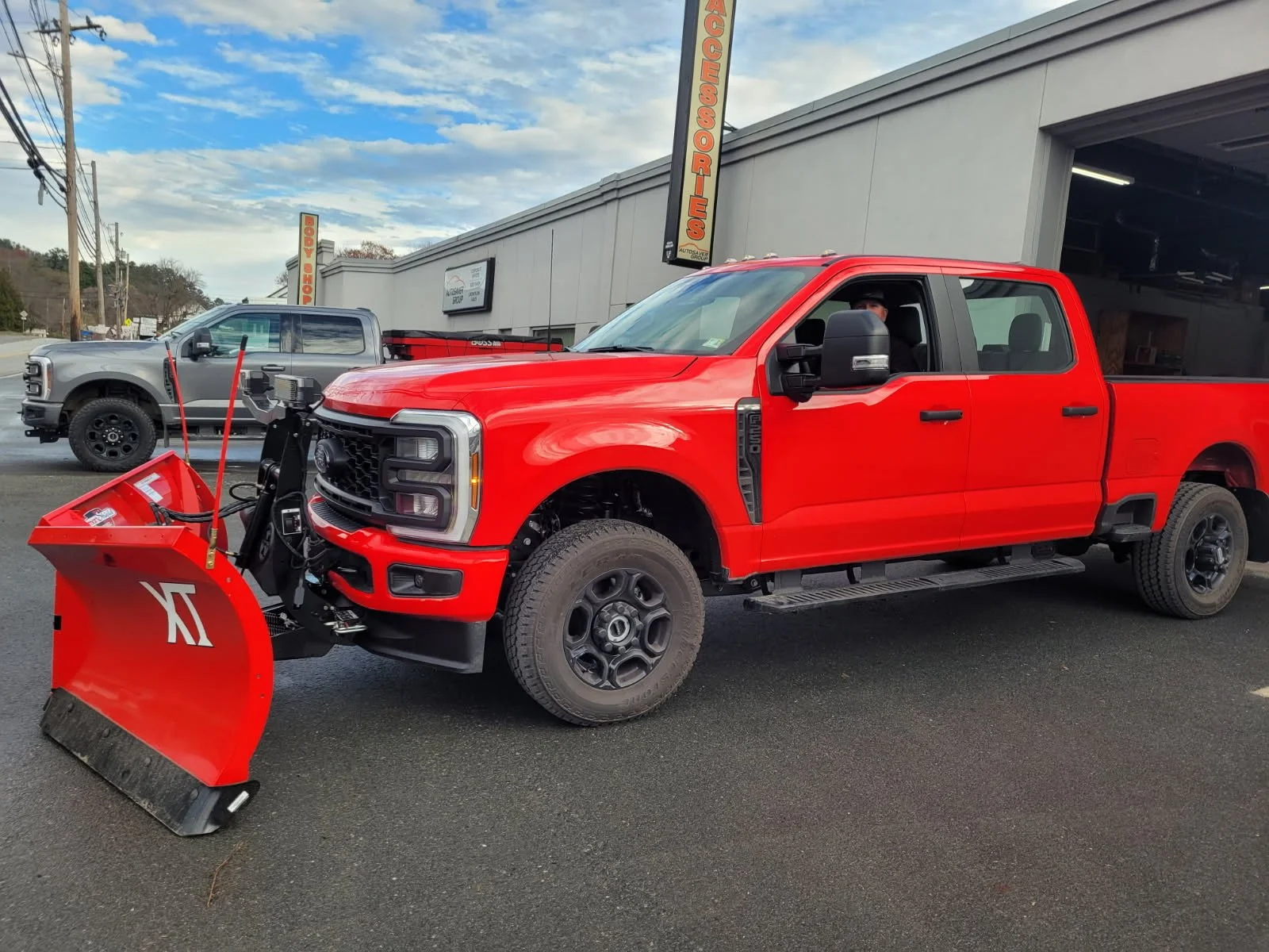 Red pickup truck with snow plow attachment parked in front of an auto accessories store, with a gray pickup truck parked nearby, under a partly cloudy sky.