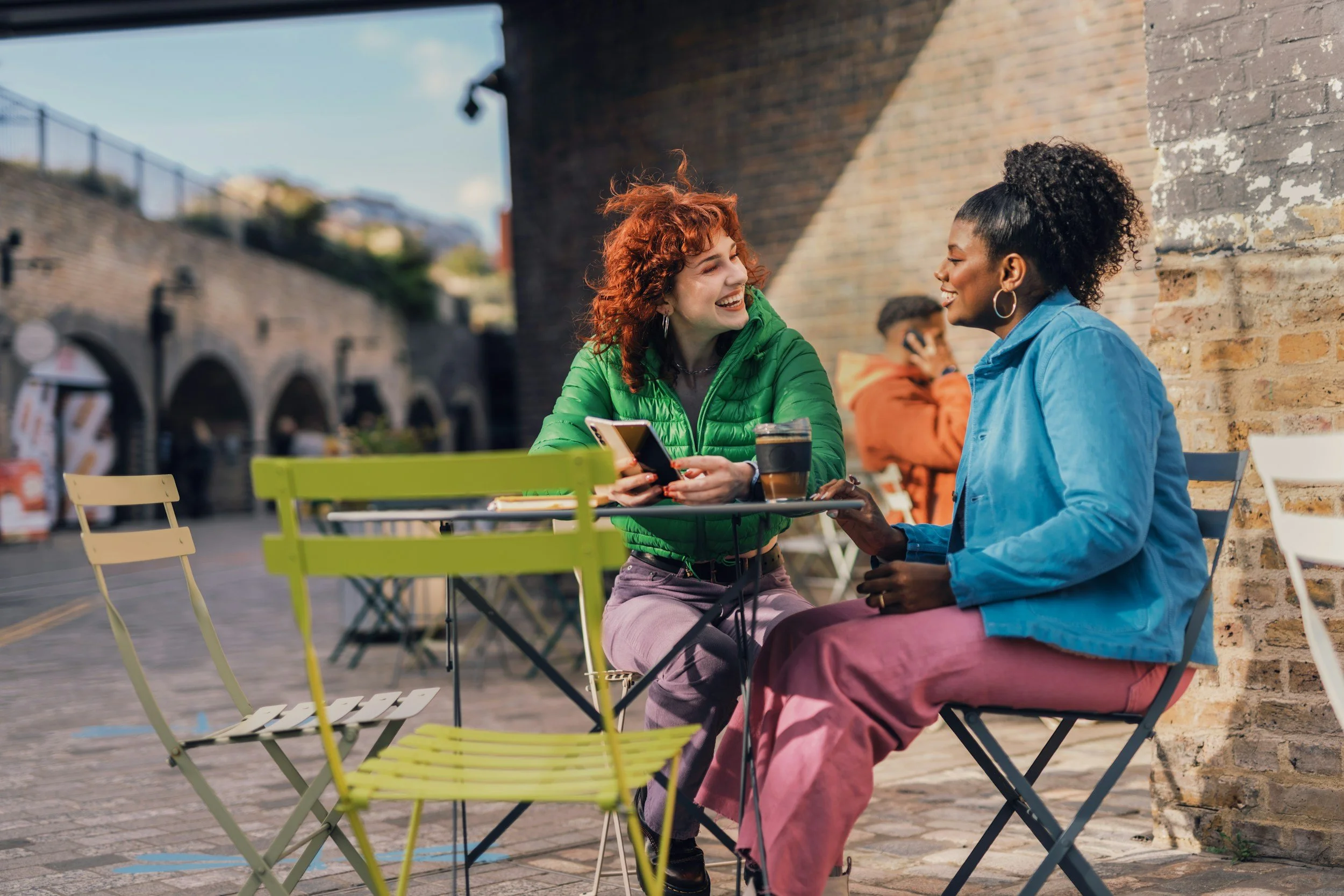 Two women sitting at an outdoor cafe table, smiling and talking, with a man in the background talking on his phone.