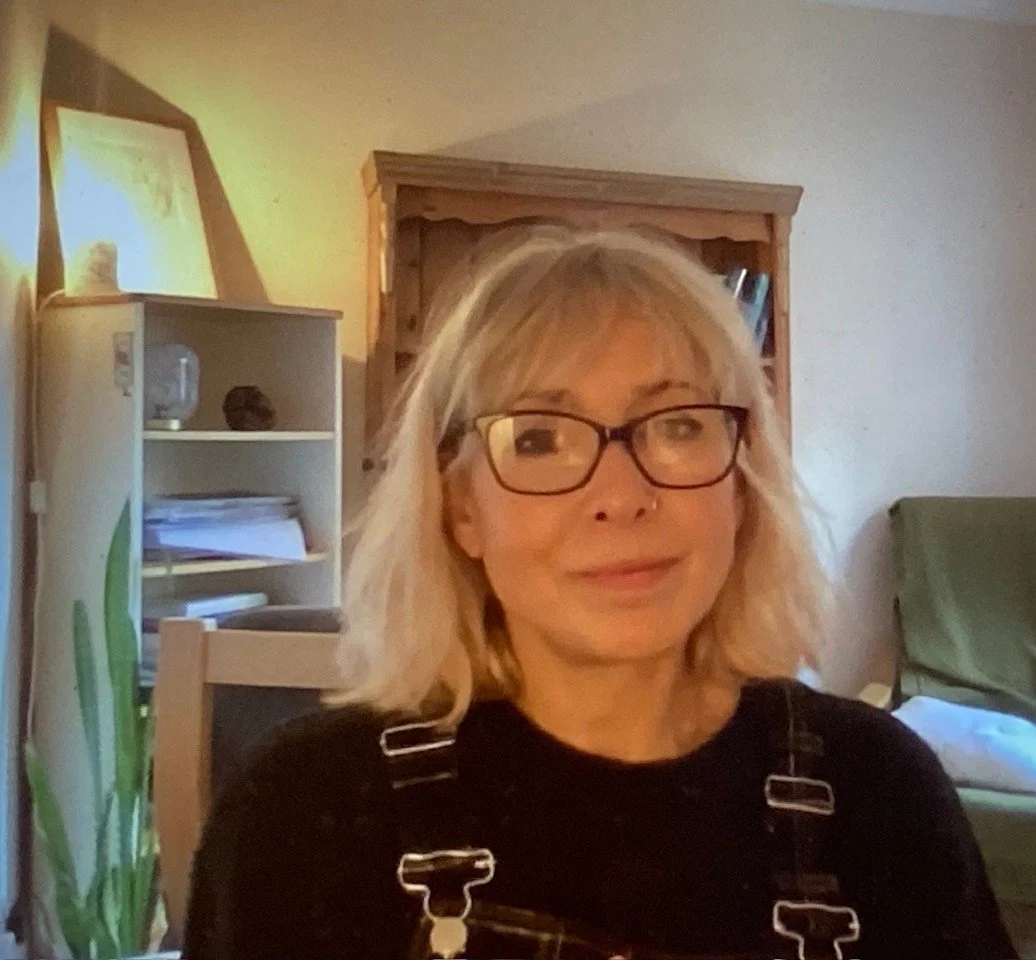 A woman with blonde hair, glasses, and a nose piercing, smiling in a cozy home setting.