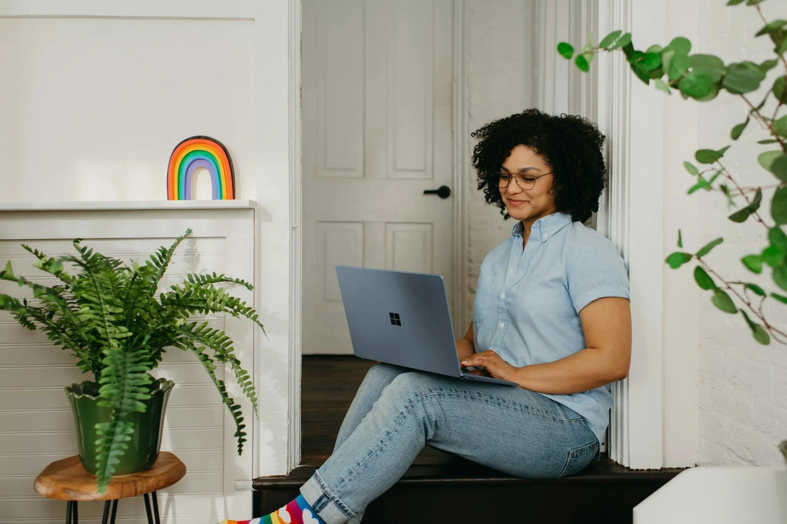 A woman with curly hair and glasses sitting on a staircase, using a Microsoft Surface laptop, with houseplants and a rainbow decor in the background.