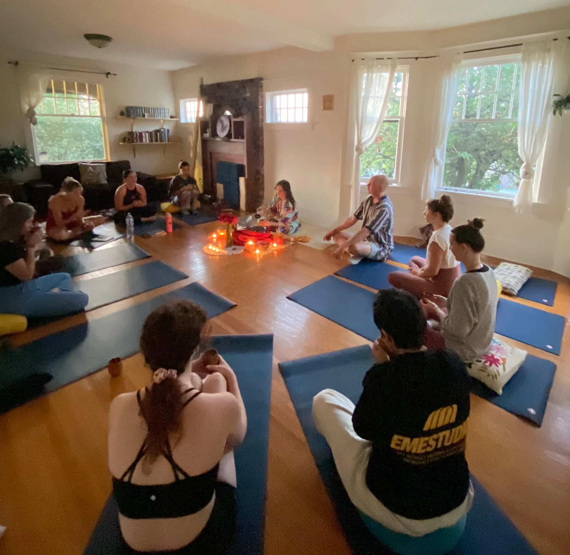 People participating in a meditation or yoga session in a living room, seated on mats around a central area with candles and decorative objects.