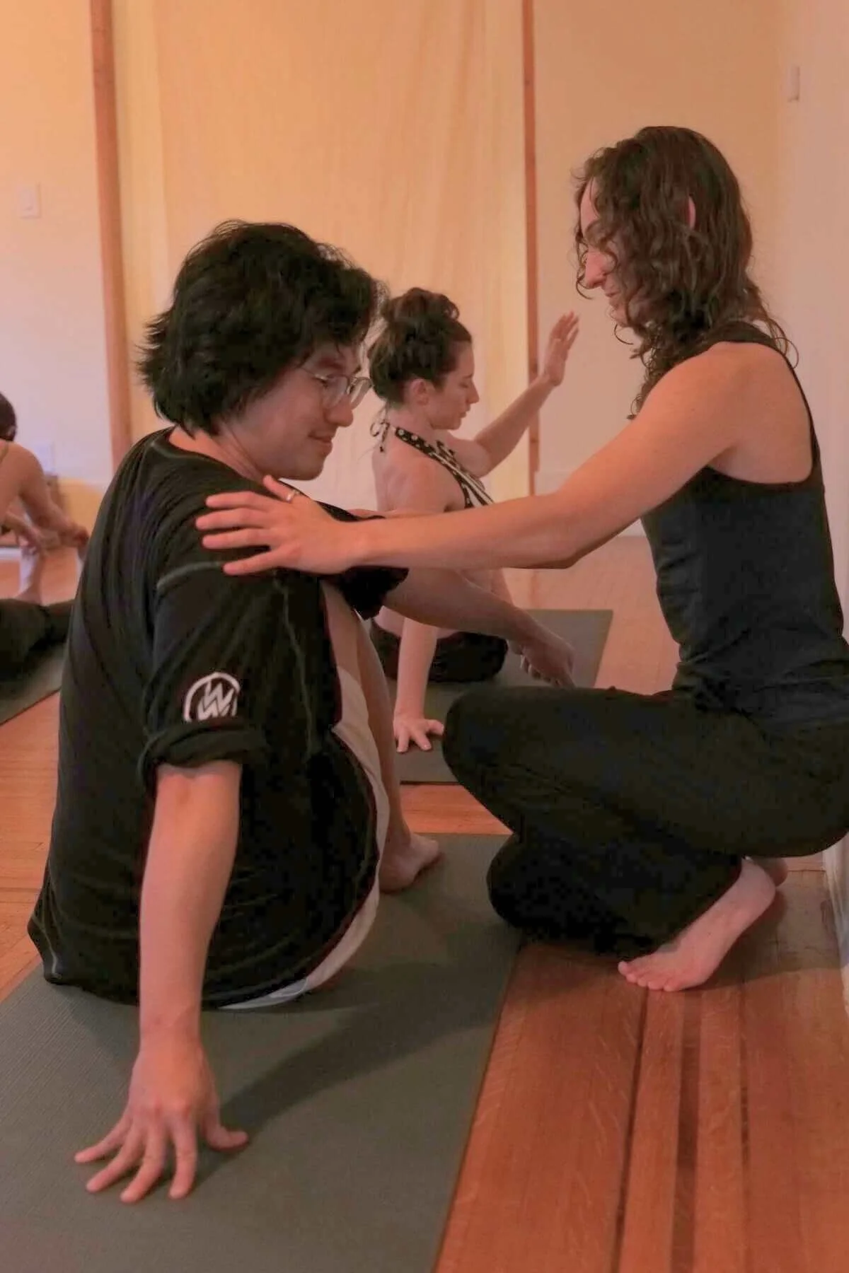 A yoga teacher is teaching headstands or a similar activity with a man sitting on the floor, while a woman in the background raises her hand in a yoga pose.