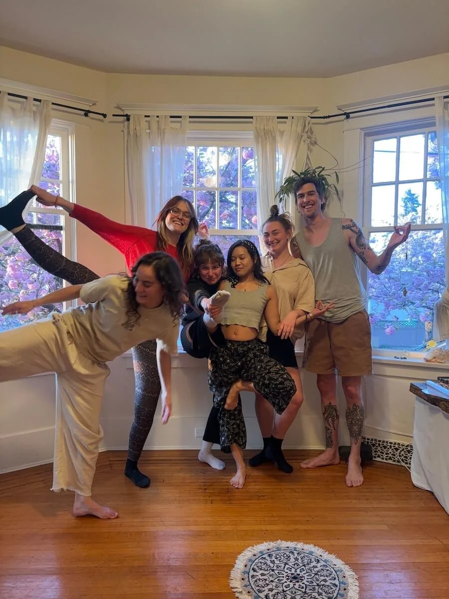 A yoga class posing playfully in a bright room with large windows and sheer curtains, with trees visible outside.