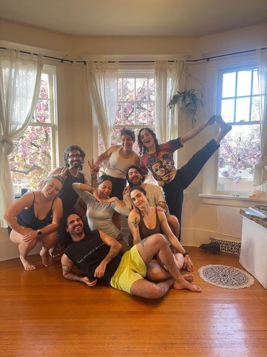 Yoga class in a bright room with large windows and white curtains, posing happily for a photo, with one person doing a high kick.