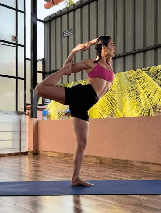 Rae roots yoga practicing yoga outdoors on a mat, performing a dancer's pose with one leg bent behind her and grasped by her hand, while the other leg supports her.