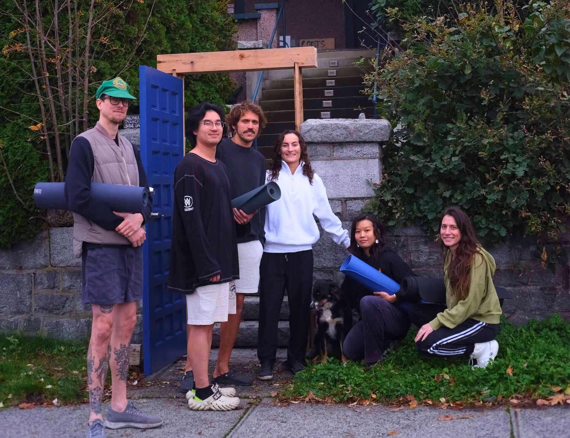 Yoga group class outside a yoga studio shala with outdoor clothing holding yoga mats, standing and sitting near a stone wall, with greenery in the background.