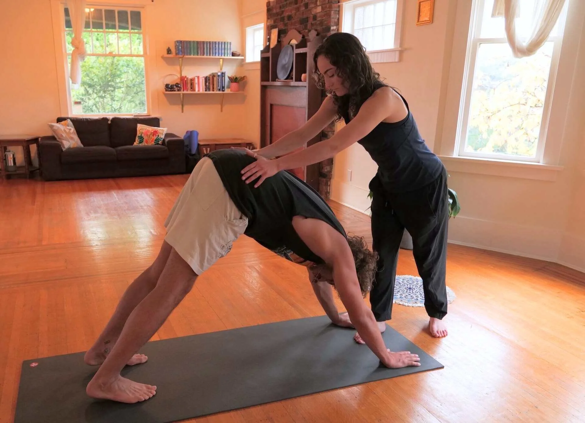 A yoga teacher assisting a man in a downward dog yoga pose inside a living room with wooden floors, a black couch, shelves with books, and large windows letting in natural light.