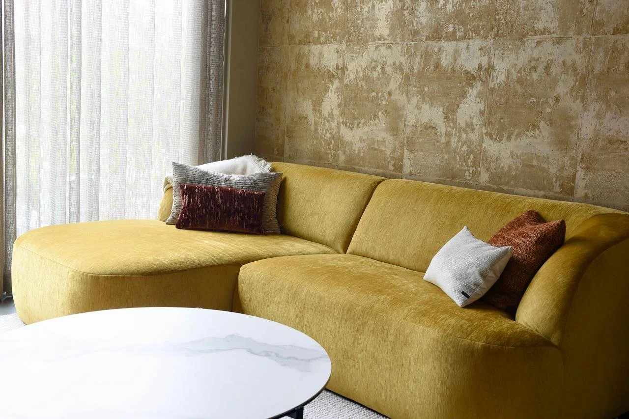 A mustard yellow sofa with multiple decorative pillows in a living room with a textured beige wall and sheer curtains.